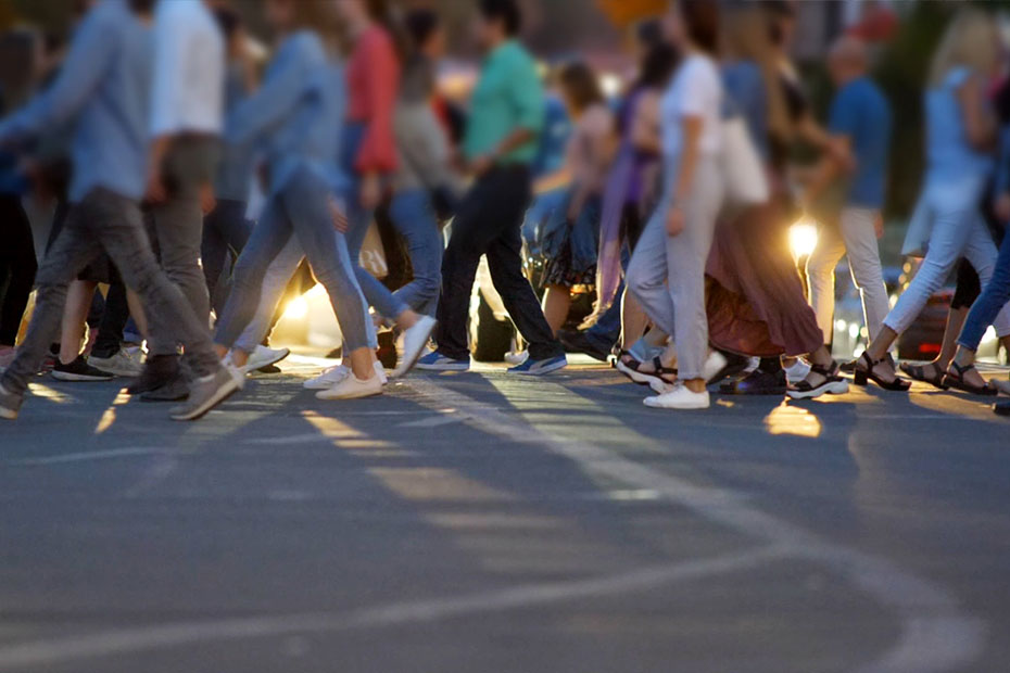 Auf dem Bild ist eine Menschenmenge zu sehen, die bei Dämmerung oder in der frühen Nacht über einen Zebrastreifen geht. Etwa ein Dutzend Personen bewegen sich gleichzeitig von links nach rechts. Viele tragen helle Sommerkleidung wie Hosen, Röcke und leichte Oberteile; überwiegend helle Turnschuhe und Sandalen sind sichtbar. Die Gesichter sind unscharf und nicht erkennbar. Im Hintergrund leuchten die Scheinwerfer wartender Autos, die die Szene zusätzlich erhellen. Der Fokus liegt auf den Beinen und Füßen der Passanten, wodurch die Bewegung besonders betont wird.